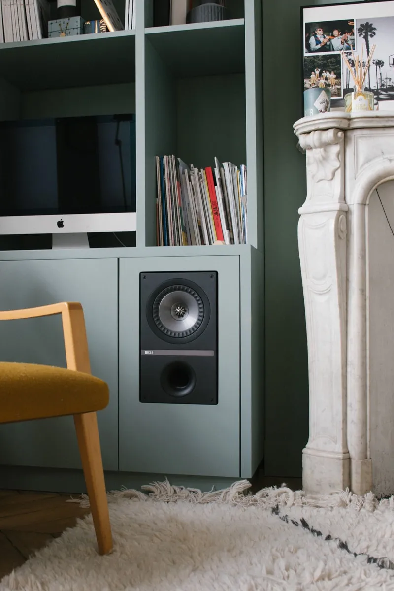 Bibliothèque sur mesure vert sauge avec enceinte encastrée, près d’une cheminée en marbre blanc, fauteuil moutarde et tapis berbère