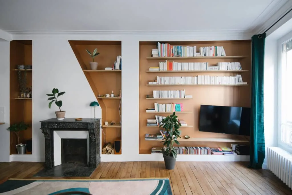 Salon rénové avec parquet ancien, cheminée en marbre noir et niches sur mesure à fond ocre dont une grande bibliothèque murale intégrant un téléviseur