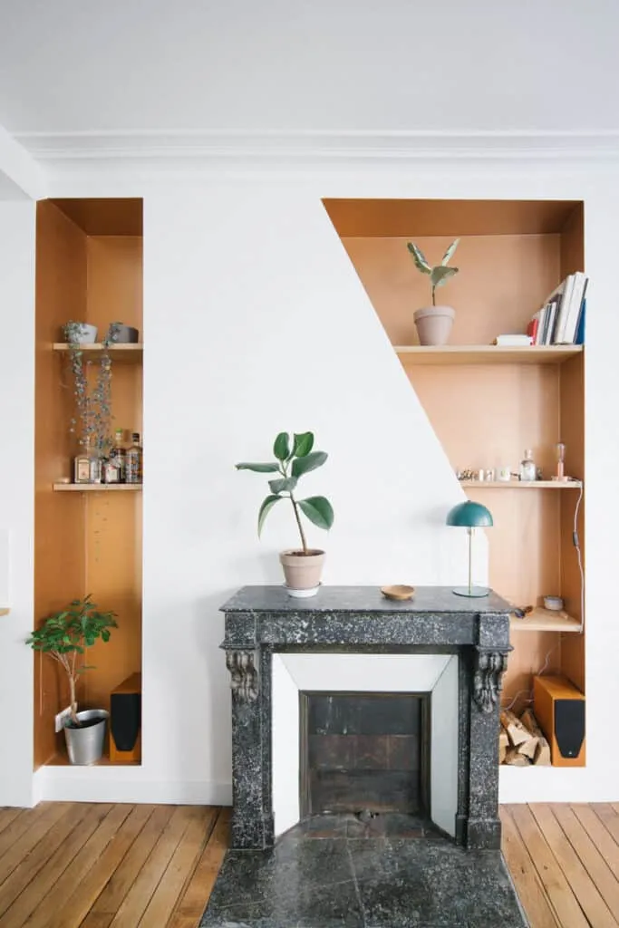 Cheminée en marbre noir entourée de niches murales sur mesure aux fonds orangés, avec plantes et étagères en bois