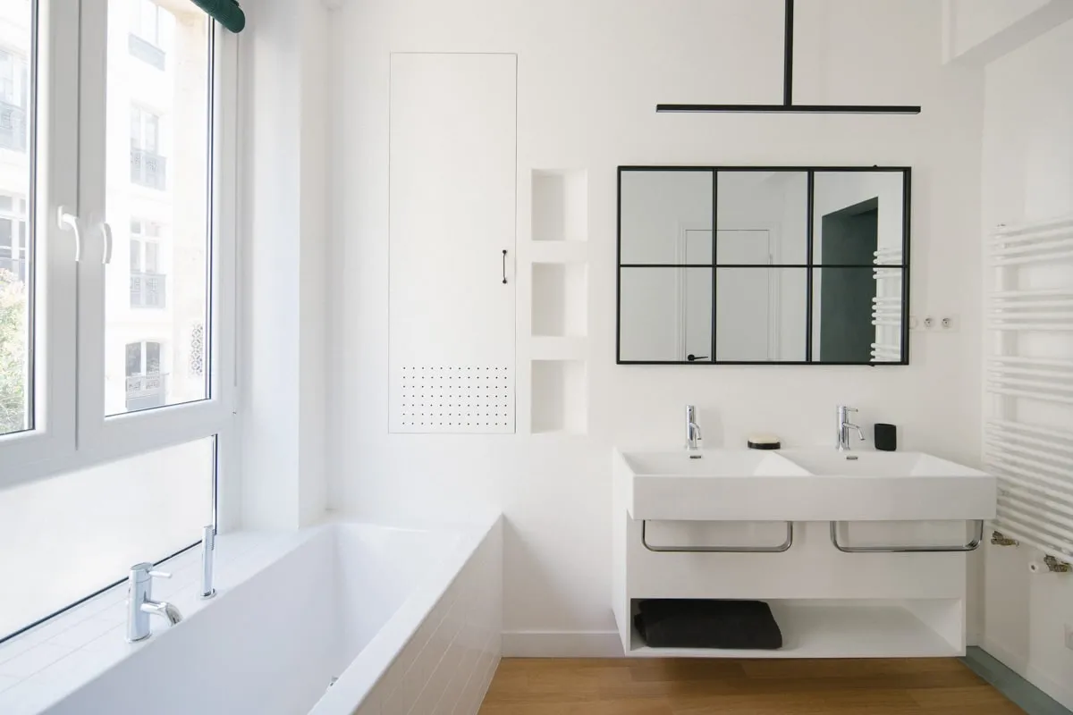 Salle de bain rénovée toute en blanc avec baignoire sous la fenêtre, meuble double vasque suspendu, miroir à cadre noir et parquet clair