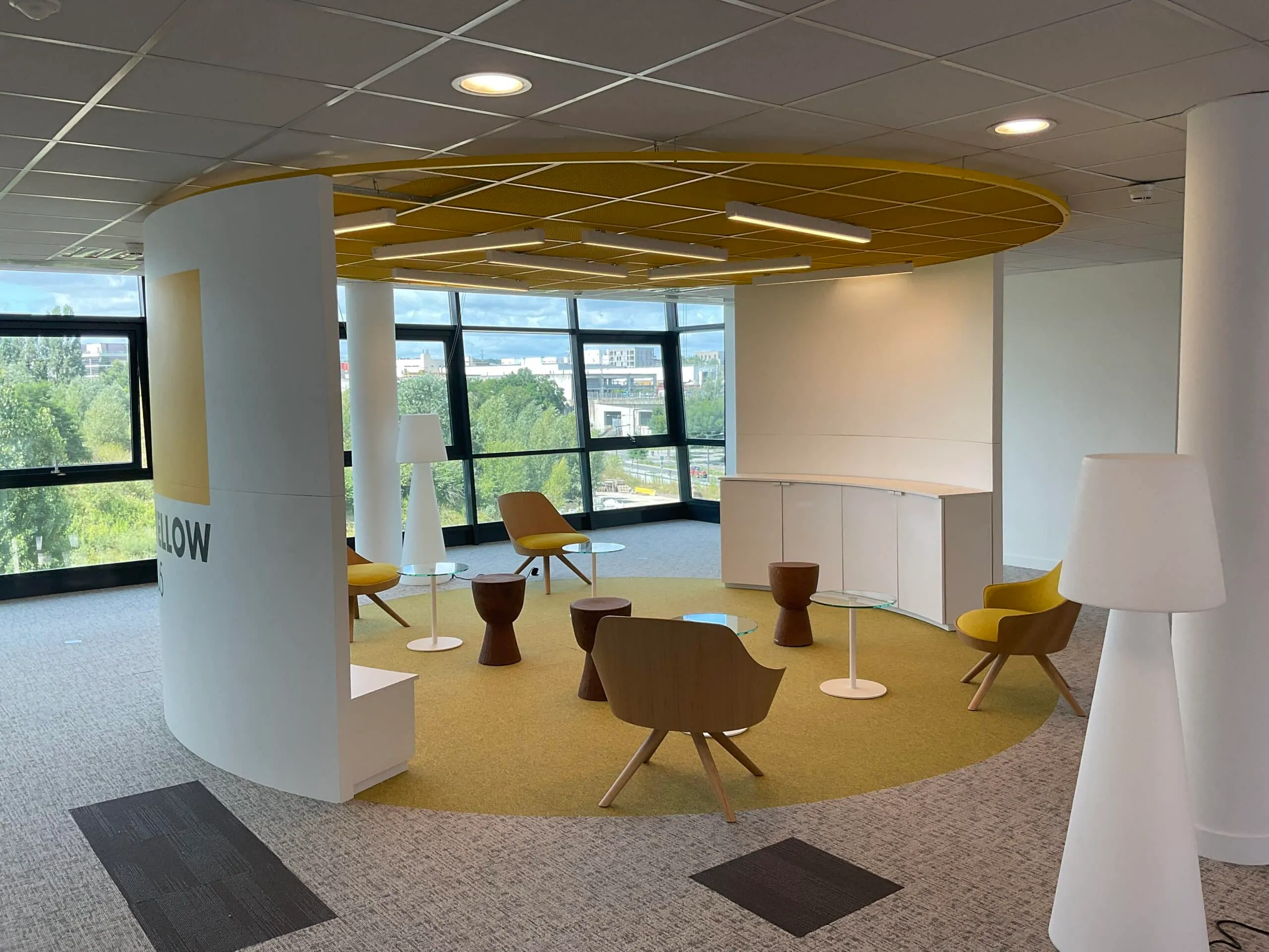 Espace de détente circulaire dans un open space de bureaux, avec cloison courbe blanche, plafond jaune, fauteuils en bois et moquette jaune
