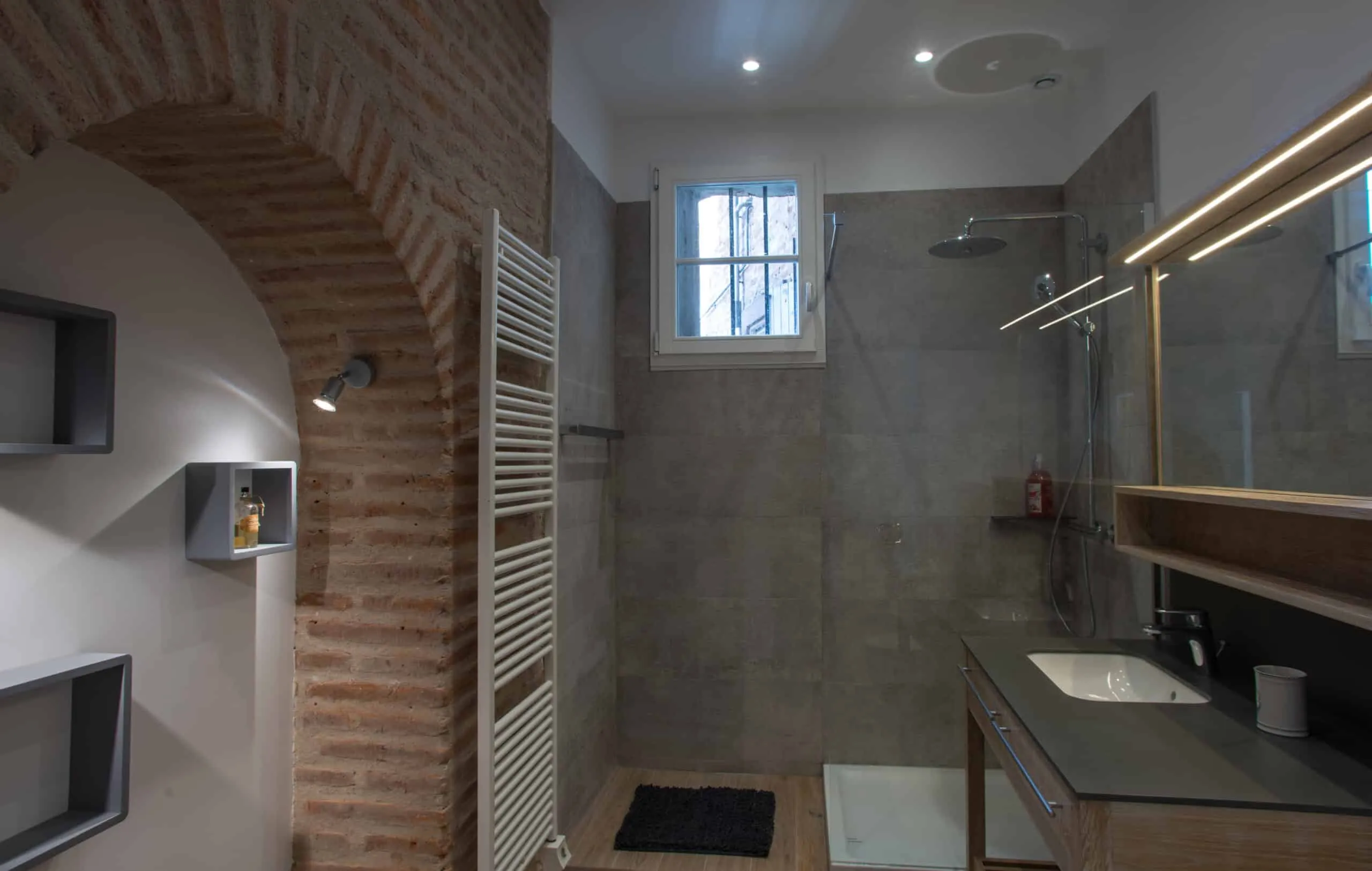 Salle de bain avec arche en briques apparentes, douche à l’italienne en carrelage gris, sèche-serviettes blanc et meuble vasque en bois avec miroir lumineux