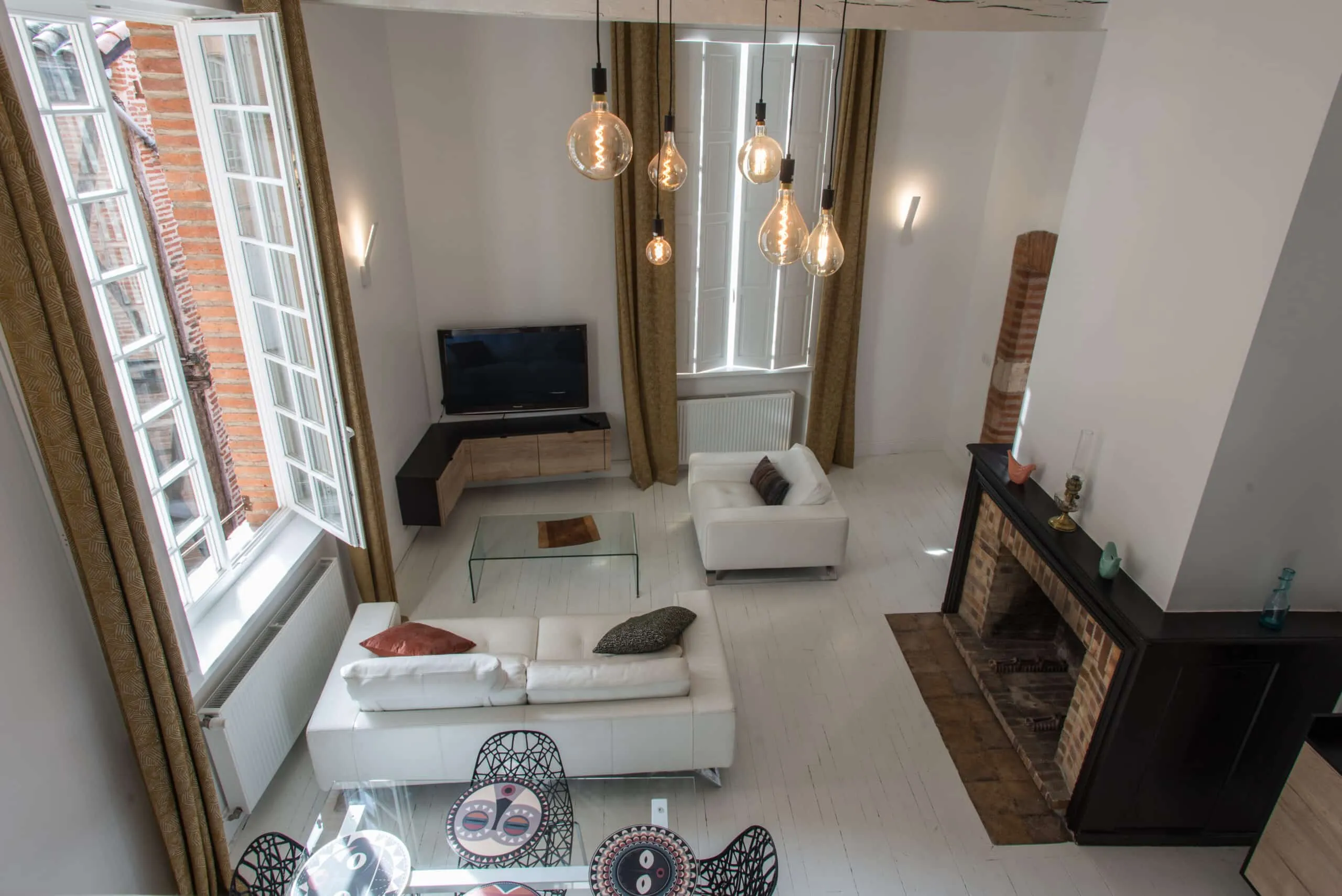 Vue en plongée d’un salon rénové au parquet blanc, avec canapés en cuir blanc, cheminée en briques et meuble TV d’angle sur mesure