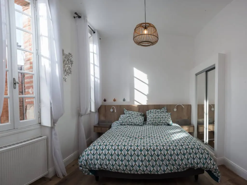 Chambre claire avec grandes fenêtres, tête de lit en bois et suspension en rotin