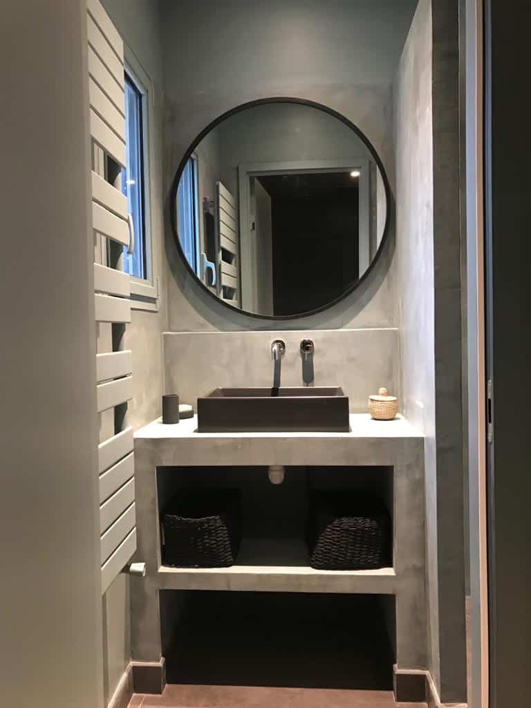 Salle d'eau contemporaine avec meuble vasque en béton ciré, lavabo carré brun et miroir rond encadré de noir. Chauffage serviette blanc.