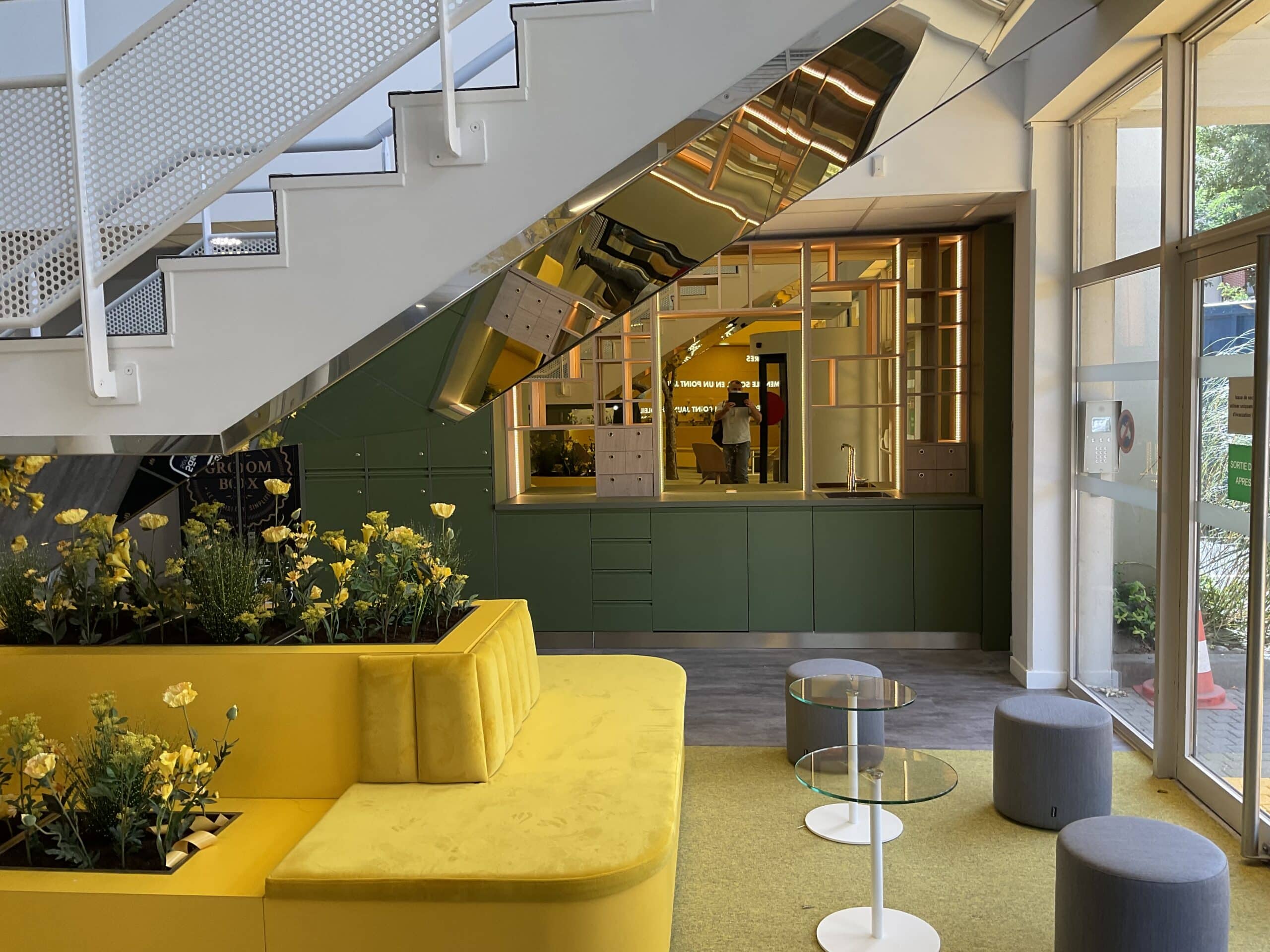 Espace d'accueil moderne avec canapé jaune vif, plantes à fleurs jaunes et comptoir vert foncé. Réflection sur la rampe d'escalier chromée. Texte visible : "GROOM BOX".