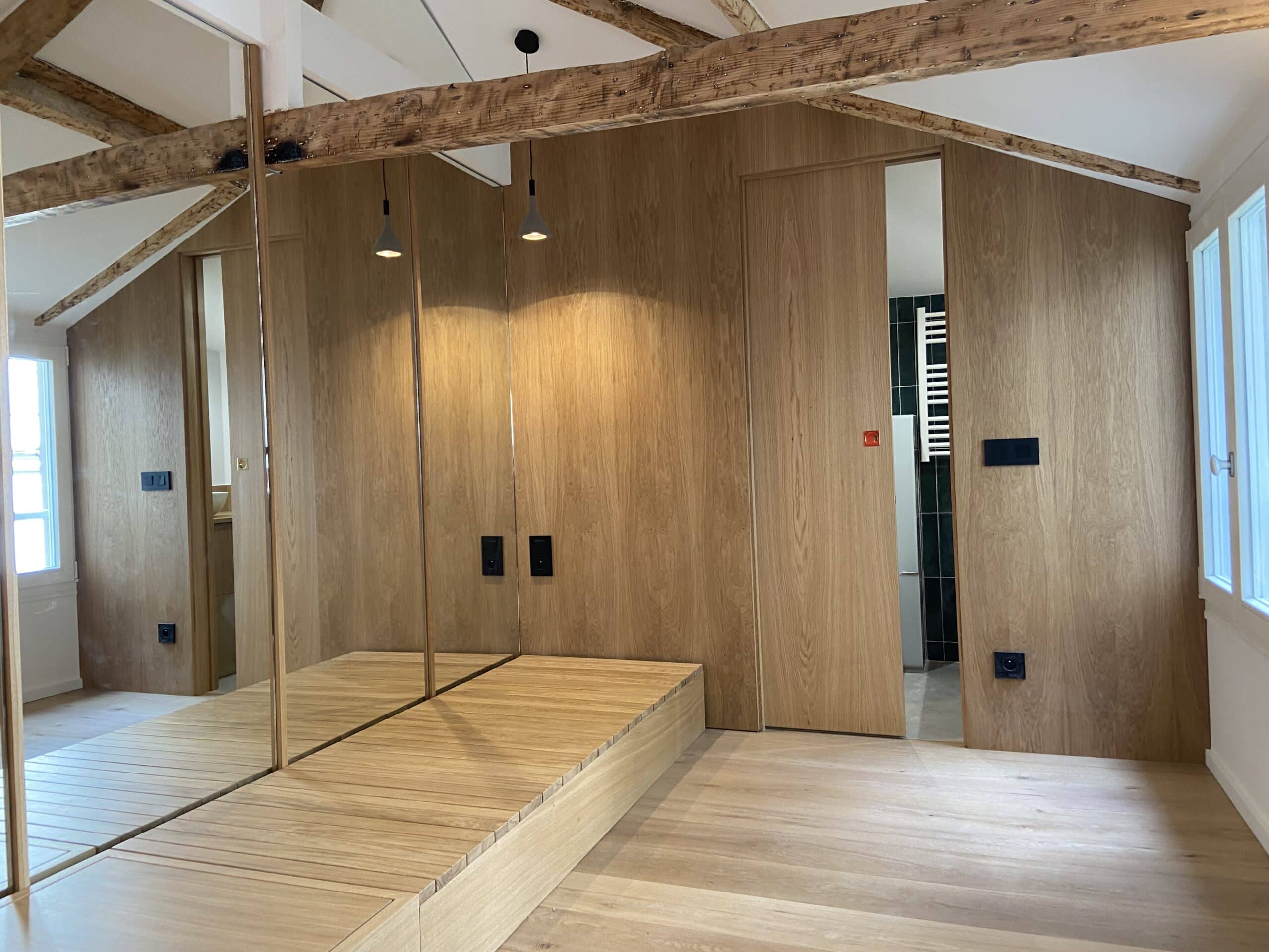 Chambre mansardée avec poutres apparentes, murs et lit en bois clair, grands miroirs et porte coulissante menant à une salle de bain sombre.