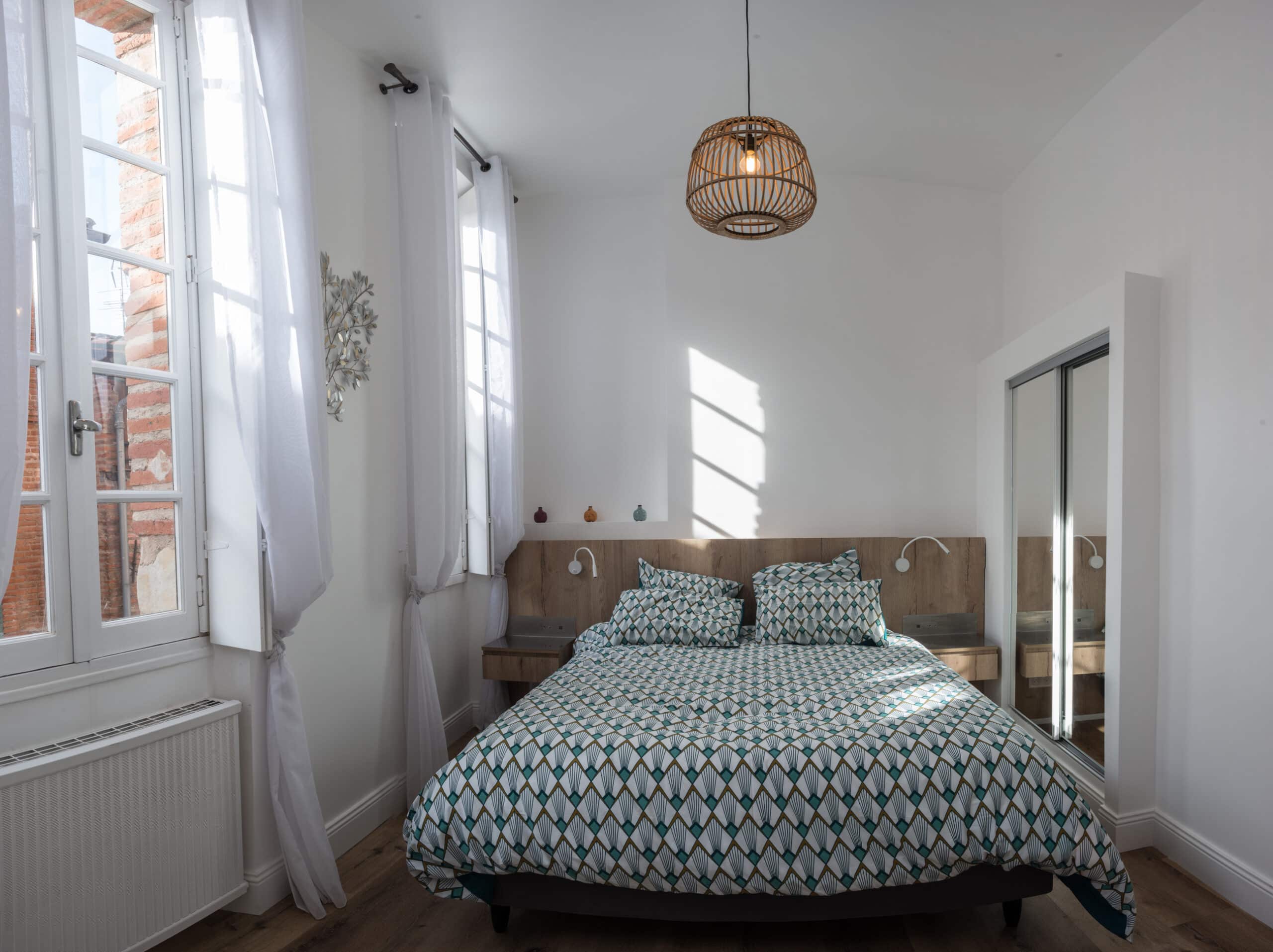 Chambre lumineuse avec lit double à motif géométrique vert et blanc, tête de lit en bois, suspension en rotin et fenêtre avec voilages blancs.