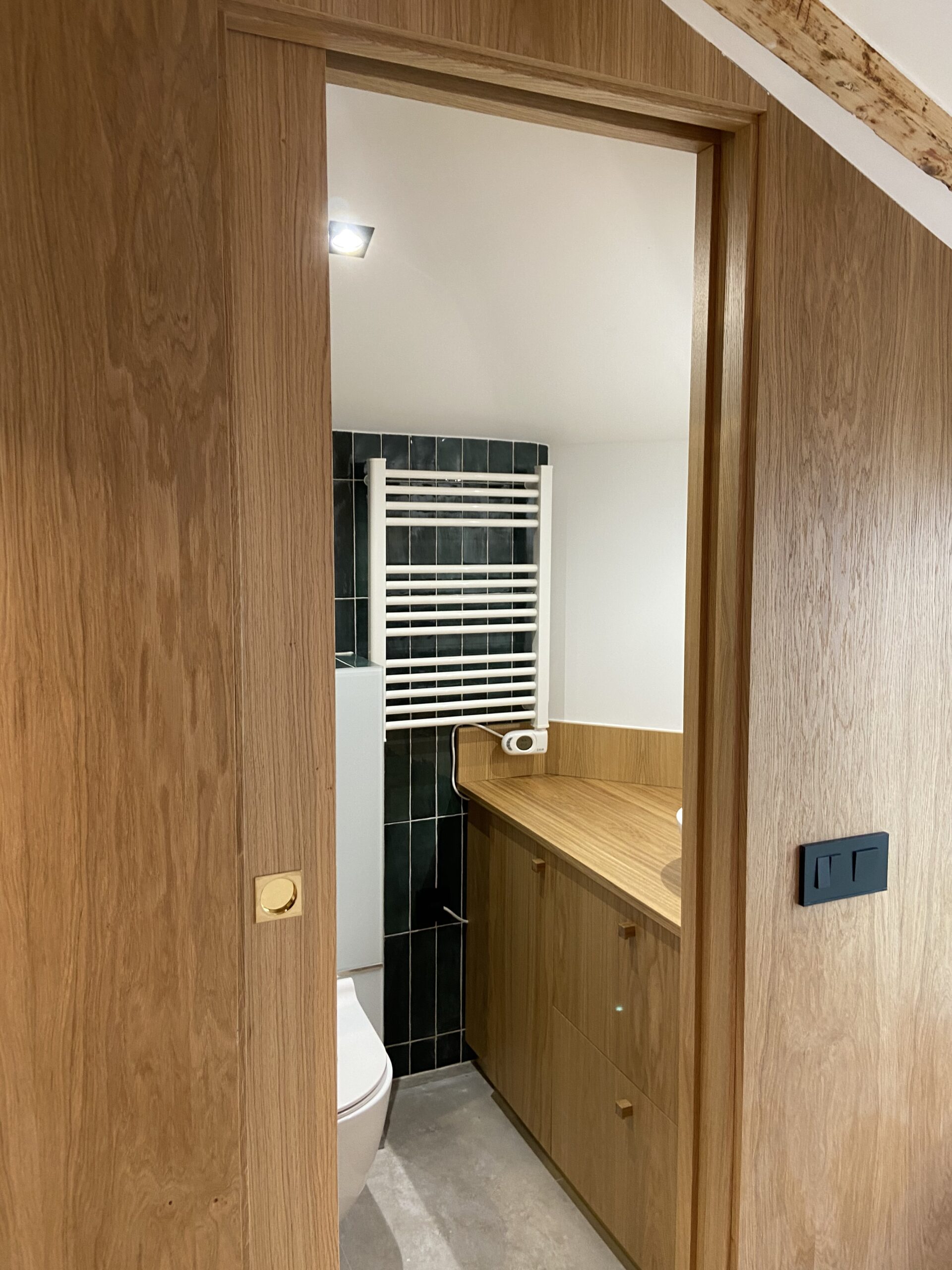 Porte coulissante en bois de chêne ouvrant sur une salle de bain moderne avec carrelage vert émeraude et sèche-serviettes blanc.