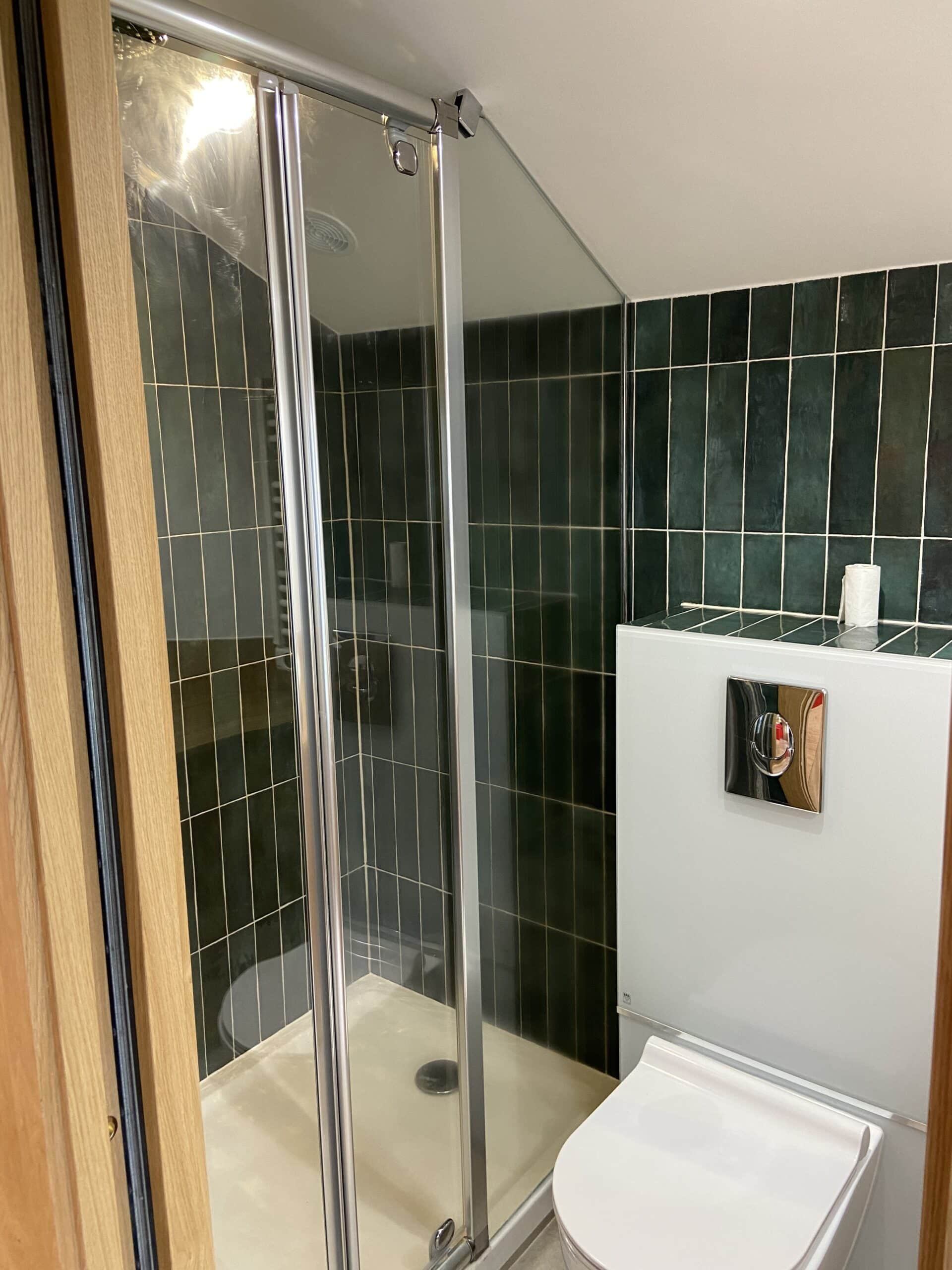 Salle de bain moderne avec douche carrelée vert forêt, WC suspendu avec plaque de commande chromée, et porte de douche vitrée.