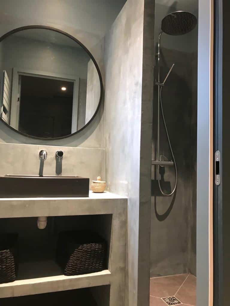 Salle de bain moderne avec murs en béton ciré, vasque rectangulaire noire, miroir rond encadré et douche à l'italienne